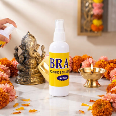 A 100ml brass polishing spray bottle next to a polished brass diya on a marble surface with burnt sienna and rose floral deco